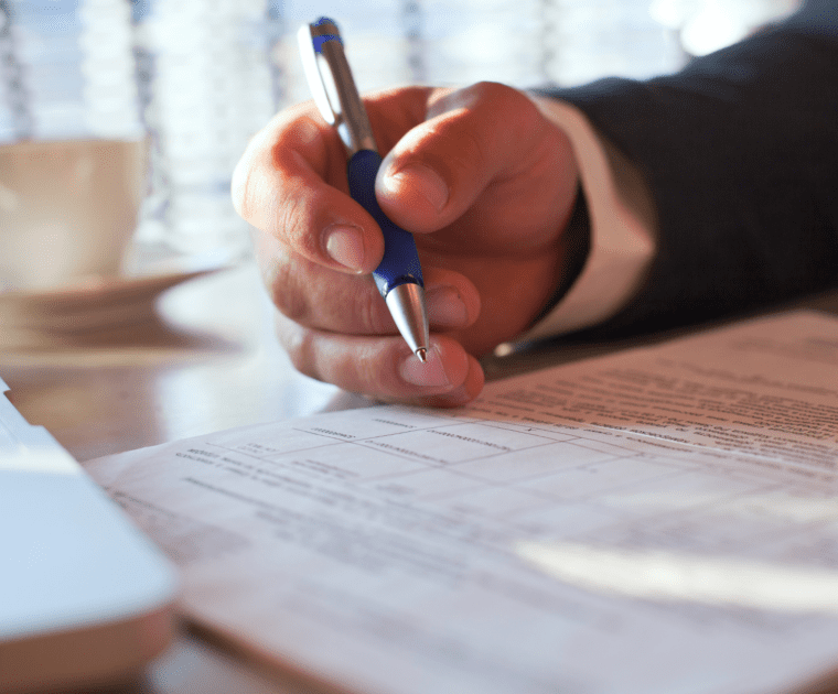 Person signing a regulatory document with a pen on a desk next to a laptop.